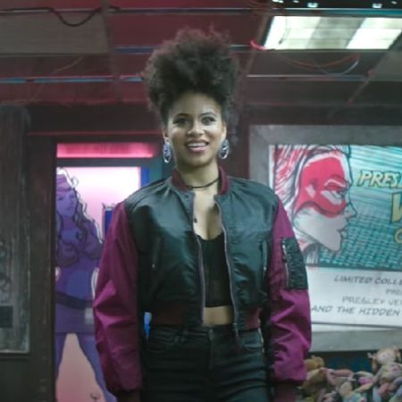Domino is wearing a black jacket with purple sleeves and has short curly hair.